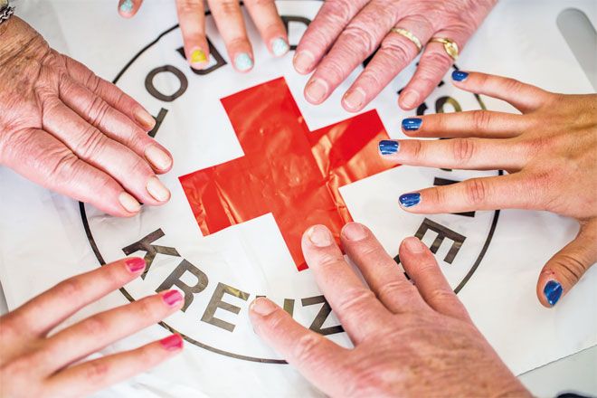 Foto: Sechs Menschen stehen im Kreis und halten jeweils eine Hand über das große DRK-Logo, das in der Mitte auf dem Tisch liegt. Zu sehen sind nur die Hände und das Logo. Es scheint, als wollten sie alle nach dem Roten Kreuz greifen.